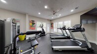 This single-floor fitness center in Union City, GA, is a well-equipped exercise facility designed to support a comprehensive workout routine. The space is thoughtfully laid out to accommodate both cardio and strength training activities. Multiple Spirit treadmills are positioned along one side of the room, offering users a dedicated area for aerobic exercise, while an elliptical machine provides an additional low-impact cardio option. The strength training section features a cable machine system and a leg press/extension machine, giving users access to a range of resistance exercises. Durable rubber-speckled flooring covers the entire space, providing a safe and resilient surface for all workout activities. Natural light streams in abundantly through numerous windows fitted with white horizontal blinds, creating a bright and energizing atmosphere. Recessed lighting and a ceiling fan overhead ensure proper illumination and airflow throughout the facility. The walls are adorned with vibrant, large-format sports-themed artwork and colorful abstract prints, adding visual energy to the environment. A wall-mounted flat-screen television, a small glass-top desk, and a sanitizing station are also present, adding practical convenience. The neutral-toned walls and clean interior finishes contribute to a professional and welcoming fitness environment.