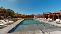 Located at 339 Boston Post Rd E in Marlborough, Massachusetts, this generously sized outdoor amenity space offers a resort-style experience within a residential community. The centerpiece of the area is a large, rectangular swimming pool with crystal-clear water and lane markers, finished with sleek tile detailing and accessible via stainless steel entry rails. The expansive surrounding deck is paved with neutral-toned stone tiles, providing ample space for relaxation and recreation. A striking wood pergola anchors one end of the pool deck, offering a shaded lounge area outfitted with comfortable seating, including cushioned chairs and chaise lounges ideal for unwinding. Additional sun loungers are arranged along the pool's perimeter, maximizing opportunities for outdoor enjoyment. The space is enclosed by a black iron security fence, ensuring privacy and safety. An auxiliary building is situated nearby, likely serving as a pool house or utility structure. The backdrop features mature trees displaying vibrant autumn foliage in shades of orange, red, and gold, creating a picturesque seasonal setting. The multi-story residential building adjacent to the pool showcases a modern exterior combining stone veneer and horizontal siding, adding an architectural sophistication to the overall environment. Tall light poles ensure the area remains functional into the evening hours.