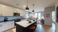 This thoughtfully designed single-floor residence seamlessly blends modern functionality with contemporary style. The open-concept layout connects the kitchen and living area, creating a bright and cohesive space ideal for both everyday living and entertaining. The kitchen is a standout feature, showcasing two-tone cabinetry with crisp white uppers and dark lower cabinets, a light-veined quartz waterfall island with bar seating, dark countertops, a light blue subway tile backsplash, and a full suite of stainless steel appliances. Stylish matte black pendant lights hang above the island, adding a modern accent. The living area flows naturally from the kitchen, bathed in natural light from large windows and a glass door that opens to the outdoors. The bedroom is a serene retreat with wide windows, warm wood-toned flooring, a ceiling fan, and ample space for a king-sized bed. The full bathroom features a soaking tub with a white subway tile surround and dark tile flooring. Conveniently adjacent, a stacked in-unit washer and dryer sits beside a spacious walk-in closet with wire shelving. Warm wood-look flooring runs throughout, tying the entire residence together with a cohesive, welcoming ambiance.