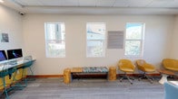 This business center is a thoughtfully designed, single-floor workspace that blends functionality with a welcoming aesthetic. The space is organized into distinct zones, each serving a specific purpose while maintaining a cohesive, contemporary feel throughout.

The main area features a large wooden conference table surrounded by blue rolling chairs, ideal for meetings or collaborative work. A wall-mounted flat-screen display anchors one end of the room, complemented by a soft teal accent wall that adds a touch of color to the otherwise neutral palette of whites and grays.

Adjacent to the conference area is a dedicated computer workstation zone, outfitted with multiple desktop computers—including both PC and Mac units—arranged along a wall-mounted counter with a yellow fascia. Teal wire-frame bar stools provide seating, and a laser printer is conveniently stationed at one end of the counter.

A comfortable lounge area with yellow swivel chairs, a rustic wooden bench supported by natural log legs, and a small side table offers a relaxed setting for informal use or waiting. A posted Business Center Policies sign is visible nearby.

Natural light filters in through several windows overlooking the surrounding neighborhood, enhancing the bright and open atmosphere. Patterned carpet tiles cover the floor throughout, tying the space together with a clean, professional finish.