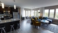 This stylish, single-floor space showcases an open-concept layout that seamlessly integrates the kitchen, dining, and living areas into one cohesive environment. The kitchen features dark cabinetry, a stainless steel refrigerator, a granite-topped island with bar seating, and pendant lighting — offering both functionality and aesthetic appeal. Adjacent to the kitchen, the dining area provides an intimate setting with a round table and upholstered chairs, positioned near expansive windows that flood the space with natural light and frame views of the surrounding greenery. The living area is anchored by a deep charcoal sectional sofa accented with bold yellow and patterned throw pillows, complemented by a pair of striking mustard-yellow armchairs that add warmth and personality. Track lighting runs along the ceiling, while sheer white curtains frame the large, multi-pane windows, enhancing the bright and airy atmosphere. Rich hardwood-style flooring runs throughout, unifying the space and adding a sense of warmth. The overall ambiance is modern and sophisticated, with a carefully curated palette of charcoal, grey, and yellow tones that creates a dynamic yet inviting interior.