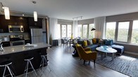 This stylish, single-floor space showcases an open-concept layout that seamlessly integrates the kitchen, dining, and living areas into one cohesive environment. The kitchen features dark cabinetry, a stainless steel refrigerator, a granite-topped island with bar seating, and pendant lighting — offering both functionality and aesthetic appeal. Adjacent to the kitchen, the dining area provides an intimate setting with a round table and upholstered chairs, positioned near expansive windows that flood the space with natural light and frame views of the surrounding greenery. The living area is anchored by a deep charcoal sectional sofa accented with bold yellow and patterned throw pillows, complemented by a pair of striking mustard-yellow armchairs that add warmth and personality. Track lighting runs along the ceiling, while sheer white curtains frame the large, multi-pane windows, enhancing the bright and airy atmosphere. Rich hardwood-style flooring runs throughout, unifying the space and adding a sense of warmth. The overall ambiance is modern and sophisticated, with a carefully curated palette of charcoal, grey, and yellow tones that creates a dynamic yet inviting interior.