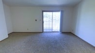 This single-floor space offers a straightforward and functional layout designed for comfortable everyday living. The main living area is finished with wall-to-wall carpet in a neutral tone, providing a soft underfoot feel that adds warmth to the room. Crisp white walls keep the space feeling clean and open, offering a versatile backdrop for a variety of interior styles.

A standout feature of the living area is the large sliding glass door framed with wood trim, which serves as both a visual focal point and a practical access point to an outdoor balcony or patio. The door allows generous amounts of natural light to pour into the room, creating a bright and airy atmosphere throughout the day. Sheer curtains flank the door, offering light diffusion while maintaining an open feel.

The room includes standard electrical outlets and wall fixtures positioned at convenient intervals. Baseboard trim runs along the perimeter of the room, adding a subtle architectural detail that frames the flooring. The overall ambiance of the space is calm and neutral, making it well-suited as a blank canvas for personalization. The connection to the outdoor area enhances the sense of space and provides an inviting extension of the interior living environment.