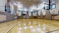 Located at 5009 Park Central Drive in Orlando, Florida, this expansive indoor sports facility offers a generously sized, single-floor gymnasium designed to accommodate serious athletic activity. The space is dominated by a gleaming, polished hardwood floor marked with multiple full and cross-court basketball configurations, allowing for simultaneous or segmented use of the floor.

Soaring ceilings supported by an open steel truss structure give the gymnasium an impressive sense of vertical volume, while rows of clerestory windows positioned high along all four walls invite abundant natural light, reducing reliance on the overhead fluorescent and pendant lighting system. The walls feature a striking geometric design, with bold triangular gray panels punctuated by the window openings, lending a modern and dynamic aesthetic to the interior.

Multiple regulation basketball hoops — both fixed and wall-mounted, adjustable units — line the perimeter, supporting multi-court play. A full electronic scoreboard is mounted prominently on one end wall for competitive game use. Retractable divider curtains allow the facility to be partitioned into separate playing zones as needed. Padded wall protectors are installed at lower wall sections to enhance player safety. Multiple access doors provide convenient entry and exit points throughout the space.