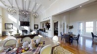 This single-story residence showcases an expansive open-concept floor plan that seamlessly connects the living, dining, and kitchen areas into one cohesive, light-filled space. The vaulted ceiling with decorative exposed beams serves as a defining architectural feature, creating a dramatic sense of height and grandeur throughout the main living area. Two statement globe chandeliers hang at varying heights, adding visual interest and warm ambient lighting.

The living area is anchored by a floor-to-ceiling stacked stone fireplace, paired with a mounted television, providing both a functional and striking focal point. Rich hardwood flooring flows throughout, unifying each zone of the open layout. The kitchen, partially visible through a framed opening, features dark cabinetry, a raised breakfast bar with industrial-style pendant lighting, and dark bar stools, offering a refined contrast to the lighter surrounding walls.

A formal dining area occupies the foreground, centered around a large wooden table with upholstered seating, while a secondary dining nook with a round table sits adjacent, offering flexible entertaining options. The overall ambiance is warm, sophisticated, and welcoming.