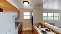 This single-floor unit offers a functional and straightforward layout with well-defined living spaces throughout. The galley-style kitchen is lined on both sides with warm wood cabinetry and granite-look countertops, providing ample storage and prep space. Equipped with a coil-burner electric cooktop, wall oven, refrigerator, dishwasher, and a stainless sink, the kitchen is fully outfitted for everyday use. A window at the end of the kitchen brings in natural light and frames views of surrounding greenery. Tile flooring completes the practical design of this space.

The open living area flows naturally from the kitchen, separated by a half-wall peninsula that adds definition without enclosing the space. Light hardwood-style flooring extends throughout the living area, enhancing the bright, airy feel created by multiple windows. A wall-mounted AC unit and baseboard heating ensure year-round comfort.

The bedrooms are carpeted and finished with clean white walls, each featuring windows that offer views of the outdoors and allow generous natural light. Baseboard heaters are present in each room. The overall ambiance of the unit is clean, neutral, and functional, offering a comfortable and adaptable living environment.