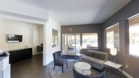 This single-floor space presents a clean, modern aesthetic defined by a thoughtful blend of neutral tones and contemporary design elements. The main lounge area serves as the centerpiece, featuring a mix of gray upholstered seating, including a sofa with patterned accent pillows, a sleek armchair with ottoman, and a glass-top coffee table, all arranged over a textured area rug atop dark hardwood flooring. Recessed ceiling lights provide even illumination throughout the space, while large windows and glass patio doors along the accent wall — finished in a deep charcoal gray — flood the interior with natural light and offer views of an outdoor seating area beyond. The contrast between the crisp white walls and the dark feature wall creates a striking visual depth. A private office or workstation nook is visible through an open doorway, providing a dedicated functional area separated from the social lounge space. The overall ambiance is polished and inviting, balancing professional utility with comfortable, hospitality-inspired design that makes the space well-suited for both relaxed gatherings and focused work.
