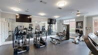 This fitness center in West Columbia, SC, offers a comprehensive exercise environment within a single, efficiently organized room. The space features durable rubber flooring throughout, crown molding, and soft neutral-gray walls that create a clean, professional atmosphere. Flush-mount ceiling lights provide consistent illumination across the entire floor.

Cardio equipment is well-represented, with multiple TRUE-brand treadmills and elliptical trainers arranged to allow comfortable use. A full dumbbell rack stocked with a wide range of free weights supports strength training, while several cable and weight machines provide options for targeted resistance workouts. A stationary bike rounds out the equipment selection.

Large windows along one wall invite natural light and offer views of the surrounding outdoor area, including a glimpse of a pool and adjacent brick buildings. Floor-to-ceiling mirrors line portions of the walls, visually expanding the space and allowing users to monitor their form during workouts. Multiple flat-screen TVs are mounted throughout, keeping members entertained during sessions.

Additional amenities include wall-mounted hooks for personal items, a waste bin, and motivational artwork. The overall design balances functionality with a polished aesthetic, making this fitness center a practical and inviting facility for daily exercise.