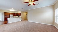 This single-floor unit is designed with a practical, open layout that connects the living area directly to the kitchen. The living room is generously proportioned, featuring warm beige walls, plush carpeting, crown molding, and a ceiling fan with a light fixture. Two windows along the exterior wall allow natural light to filter in, offering views of the surrounding exterior. A front entry door with a wood-toned tile threshold provides a clean transition into the carpeted main space.

The kitchen is outfitted with rich, dark cherry wood cabinetry extending to the ceiling, granite-style countertops, stainless steel appliances including a refrigerator, range, and over-the-range microwave, and hardwood-style flooring. A peninsula counter connects the kitchen to the living area, facilitating an open flow.

The bedrooms are carpeted and equipped with ceiling fans and window blinds. One bedroom features a spacious walk-in closet with wire shelving on multiple walls for organized storage. The bathroom includes tile flooring, a rich wood vanity, and wall-mounted grab bars, adding both function and accessibility to the space.