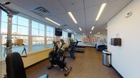 Located at 15 Poplar St in Glassboro, NJ, this dedicated fitness center offers a well-equipped, single-floor workout facility designed to support a wide range of exercise routines. The space features a drop ceiling with recessed linear LED lighting that provides bright, even illumination throughout, complemented by large windows along multiple walls that invite abundant natural light and views of the surrounding area.

The room is outfitted with a comprehensive selection of cardio equipment, including Star Trac treadmills, stationary and air resistance bikes, elliptical machines, and a rowing-style cross trainer. A multi-station cable strength machine provides options for resistance and weight training, while an assortment of medicine balls, stability balls, resistance bands, and exercise mats caters to functional and flexibility training. Wall-mounted flat-screen televisions are positioned throughout for entertainment during workouts.

A bold "FITNESS" typographic mural adorns one wall, adding an energetic and motivational aesthetic to the space. A water fountain station is conveniently located near the entrance, alongside a sanitation dispenser and waste receptacle, promoting hygiene throughout the facility. The durable carpet flooring provides comfort underfoot, and the clean, open layout ensures sufficient space between equipment for safe and comfortable use. This fitness center is well-suited for a variety of users seeking a fully equipped workout environment.