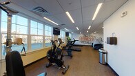 Located at 15 Poplar St in Glassboro, NJ, this dedicated fitness center offers a well-equipped, single-floor workout facility designed to support a wide range of exercise routines. The space features a drop ceiling with recessed linear LED lighting that provides bright, even illumination throughout, complemented by large windows along multiple walls that invite abundant natural light and views of the surrounding area.

The room is outfitted with a comprehensive selection of cardio equipment, including Star Trac treadmills, stationary and air resistance bikes, elliptical machines, and a rowing-style cross trainer. A multi-station cable strength machine provides options for resistance and weight training, while an assortment of medicine balls, stability balls, resistance bands, and exercise mats caters to functional and flexibility training. Wall-mounted flat-screen televisions are positioned throughout for entertainment during workouts.

A bold "FITNESS" typographic mural adorns one wall, adding an energetic and motivational aesthetic to the space. A water fountain station is conveniently located near the entrance, alongside a sanitation dispenser and waste receptacle, promoting hygiene throughout the facility. The durable carpet flooring provides comfort underfoot, and the clean, open layout ensures sufficient space between equipment for safe and comfortable use. This fitness center is well-suited for a variety of users seeking a fully equipped workout environment.