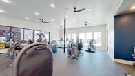 This fitness center offers a spacious, single-floor layout designed to accommodate a comprehensive range of workout activities. The space is flooded with natural light through large, multi-pane windows along the exterior walls, providing open views of the surrounding area and creating an energizing atmosphere throughout.

The flooring transitions between durable rubber and carpet surfaces, each suited to different workout zones. The cardio area features multiple treadmills, elliptical trainers, recumbent bikes, and stair climbers, all arranged to take advantage of the natural light and outdoor views. A dedicated strength training zone houses Hoist Fitness selectorized machines and a full dumbbell rack with an adjustable bench, positioned against a bold navy blue accent wall that adds visual contrast to the otherwise crisp white interior.

Full-length mirrors are mounted in the strength zone, enhancing the sense of space and supporting proper form during exercise. Multiple ceiling fans are distributed throughout, ensuring consistent airflow. Recessed lighting supplements the natural light for well-lit workouts at any hour. Structural columns in a dark finish provide architectural definition without interrupting the open floor plan. The overall design is clean, modern, and functional, making this fitness center a well-rounded facility for cardiovascular and strength training alike.