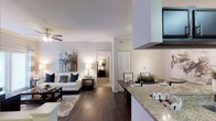 This single-story residence showcases an open-concept floor plan that integrates the kitchen, dining, and living areas into one cohesive, flowing space. Rich dark hardwood flooring extends throughout the main living area, providing a warm contrast to the light-toned walls and textured ceilings. The living room is anchored by a ceiling fan and features crown molding, lending a polished, finished look. A comfortable seating arrangement with a sofa accented by navy and neutral throw pillows sits beneath a contemporary piece of wall art, completing the inviting atmosphere.

The kitchen is positioned prominently within the open layout and features granite countertops with a raised breakfast bar, offering both additional prep space and casual seating. A large range hood serves as a functional focal point above the cooking area. Adjacent to the kitchen, the dining area provides space for a full table setup. A hallway visible from the main living space leads to additional rooms, including what appears to be a bedroom with a louvered closet door, maintaining a sense of privacy while preserving the open flow of the home.