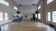 This fitness center is designed to accommodate a well-rounded workout experience within a single open floor plan. The space immediately impresses with its soaring ceilings, which are accentuated by tall, narrow clerestory windows positioned near the roofline, allowing natural light to filter in while maintaining privacy. Two ceiling fans are suspended from above, ensuring comfortable air circulation throughout the room.

The flooring consists of light hardwood-style planks, lending the space a clean and polished aesthetic. Cardio equipment, including treadmills and an elliptical trainer, is arranged along one wall, complemented by a large wall mirror that visually expands the space and allows users to monitor their form. On the opposite side, a multi-station strength-training machine and a pull-up/dip station provide options for resistance training.

Two wall-mounted flat-screen televisions are positioned at opposite ends of the room, offering entertainment during workouts. Natural light flows in through multiple windows fitted with blinds, and recessed lighting ensures consistent illumination. The neutral gray walls and open floor plan create a clean, functional, and inviting environment suitable for a variety of fitness activities.