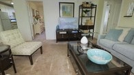 This single-floor residence offers a thoughtfully arranged and comfortably styled living environment. The living room serves as the centerpiece of the space, featuring wall-to-wall carpeting that adds warmth underfoot and a neutral color palette that creates a calm, welcoming atmosphere. A dark-toned media console anchors one wall, topped with a flat-screen television and complemented by a matching open shelving unit displaying decorative accents, woven baskets, and greenery. A glass-topped coffee table with dark framing sits at the heart of the seating area, styled with a decorative tray and a distinctive turquoise bowl that adds a pop of color. Framed artwork adorns the walls, contributing to the refined yet relaxed character of the room. A cushioned bench adds additional seating and versatility to the layout. A glimpse through an adjacent doorway reveals a secondary area furnished with a classic white dresser accented by a vase of fresh red flowers, hinting at well-maintained and consistently styled spaces throughout. The overall ambiance is clean, organized, and tastefully decorated, making this a comfortable and visually cohesive living space.