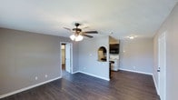 This single-floor space showcases a practical and straightforward layout designed for comfortable everyday living. The main living area features dark hardwood-style flooring that contrasts cleanly against light gray walls and white trim, creating a modern yet understated aesthetic. A ceiling fan with a built-in light fixture is centrally positioned overhead, providing both air circulation and ambient lighting to the space.

A defining architectural detail is the arched pass-through opening that connects the living area to the kitchen, adding a touch of character to an otherwise clean-lined interior. The kitchen is equipped with essential appliances, including a white range and an overhead microwave, with dark upper cabinetry providing storage and visual contrast. The pass-through also features a countertop ledge, suitable for casual dining or additional workspace.

A doorway off the living area leads to additional rooms, offering privacy and separation from the main living space. The interior is finished with clean white baseboards throughout, and the neutral color palette ensures versatility for a variety of furnishing styles. Overall, the space presents a tidy, functional environment with a cohesive design.