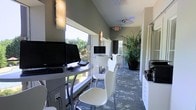 This single-floor business center offers a thoughtfully designed workspace that balances functionality with a welcoming aesthetic. The room is lined with a continuous workstation counter along the windowed wall, providing an abundance of natural light that floods the space and offers pleasant views of lush greenery outside. Multiple computer workstations, including Dell desktop setups and an iMac, are neatly arranged along the counter, making the space ideal for focused work or research. Sleek white modern chairs with chrome legs are positioned throughout, providing comfortable seating at each station. The flooring features a contemporary patterned carpet in cool gray and blue tones, adding visual interest underfoot. Vaulted ceilings with warm wall sconces create an open, airy feel, while a ceiling fan ensures comfortable airflow throughout. A tall, vibrant potted tropical plant anchors the far end of the room, introducing a natural element that softens the professional setting. Additional amenities, including what appears to be a printer or copier station along one wall, further support the space's practical, service-oriented design. The overall ambiance is clean, modern, and inviting — well-suited for a shared community or hospitality environment.