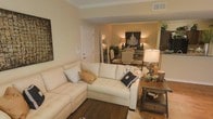 This single-story space showcases a thoughtfully designed open-concept layout that seamlessly connects the living and dining areas. The living room is anchored by a generous sectional sofa in a warm cream upholstery, complemented by decorative throw pillows and a plush area rug layered over rich hardwood-style flooring. Crown molding frames the ceiling throughout, adding a refined architectural touch to the warm, neutral color palette of soft yellows and whites.

A rustic wooden side table with a sculptural lamp adds texture and character to the living space, while framed wall art contributes visual interest. The dining area, visible through an open pass-through, features an elegant chandelier-style light fixture flanked by matching table lamps, casting a warm ambient glow over a formal dining set with upholstered chairs. Lush greenery and decorative accents further enhance the refined atmosphere. The overall ambiance of the space is warm, inviting, and well-appointed, balancing casual comfort with classic elegance throughout.