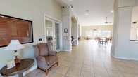 This single-story community space offers an open, airy atmosphere defined by tall ceilings and large windows that allow natural light to fill the interior. The layout flows seamlessly from a comfortable lounge area near the entrance, where a classic upholstered wingback chair provides a welcoming spot for relaxation, into a more expansive dining area beyond. Large-format ceramic tile flooring in a warm neutral tone runs throughout, lending a clean and cohesive look to the space. Architectural columns with arched detailing add visual interest and help define distinct zones within the open floor plan. French doors off the entrance lead to an adjacent room, likely used for private gatherings or activities, visible through the glass panes. The dining area is furnished with multiple tables and chairs arranged to encourage social interaction, while pendant light fixtures hang from the ceiling, complementing the recessed lighting above. The overall ambiance is calm, bright, and inviting, designed to foster community engagement in a comfortable, well-maintained environment.