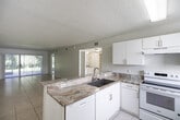 Located at 6475 Sea Wolf Ct in Naples, FL, this single-story, 2-bedroom, 2-bathroom home offers a well-organized floor plan with tile flooring throughout. The open-concept living and dining area is bright and airy, with sliding glass doors that lead to a private patio and views of the surrounding greenery. The kitchen serves as the functional heart of the home, featuring white cabinetry, striking variegated marble-look countertops, a peninsula with a stainless steel sink, and white appliances including an electric range and dishwasher. A hallway connects the main living spaces to the two bedrooms, each sized to accommodate a comfortable furnishing arrangement and equipped with closets. The primary bedroom features a large window with vertical blinds, allowing for natural light and outdoor views. The primary bathroom includes a neo-angle glass-enclosed shower with marble-style tile surround and a granite-topped vanity. The second bathroom offers a full tub-shower combination with classic white tile, complemented by a matching granite vanity. Additional spaces include a dedicated laundry area, a utility room housing the water heater with overhead wire shelving, and a hallway for convenient circulation throughout the home.