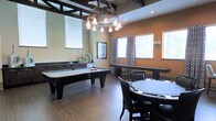 This single-floor game room is thoughtfully designed for entertainment and leisure. The space is anchored by a sleek black pool table positioned beneath a striking industrial-style chandelier, which features multiple pendant bulbs suspended from a circular metal frame, adding a bold focal point to the room. Exposed dark ceiling beams complement the chandelier, contributing to the room's contemporary yet rustic aesthetic.

Adjacent to the pool table, a round poker table surrounded by dark upholstered chairs provides a dedicated area for card games and social gatherings. The warm amber and neutral wall tones create an inviting ambiance throughout the space. Wood-look flooring runs seamlessly across the room, adding warmth and texture underfoot.

Natural light filters in generously through multiple windows dressed with white blinds and patterned curtains, brightening the interior while maintaining a sense of privacy. A low dark credenza along one wall offers functional storage and display space. Recessed lighting supplements the chandelier, ensuring the room remains well-lit. Overall, the space balances style and functionality, making it an ideal setting for recreational activities and social events.