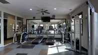 This fitness center is designed to provide a comprehensive workout environment within a compact, efficiently arranged space. The room features a lineup of cardio equipment, including multiple treadmills and an elliptical trainer, positioned to face a large wall-to-wall mirror that visually expands the space and allows users to monitor their form during exercise. A cable machine and additional strength training equipment are situated along one side, offering options for resistance-based workouts. The flooring consists of a combination of rubber and hard surface tiles, providing both durability and comfort underfoot. Overhead, a ceiling fan with a mounted flat-screen television keeps the environment comfortable and entertaining during workouts. Recessed lighting supplements the natural light that filters in through windows visible in the mirrored reflection, contributing to a bright and energizing atmosphere. The neutral gray and white color palette, combined with the mirrored wall, gives the space a clean, modern aesthetic. Overall, the fitness center is thoughtfully arranged to maximize usability, making it a practical and inviting space for daily exercise routines.