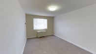 This single-floor space presents a straightforward and functional design characterized by neutral, light beige walls that create a calm and understated atmosphere. The room is finished with wall-to-wall carpeting in a soft, muted tone that complements the overall color palette and adds a layer of warmth underfoot. A single window fitted with horizontal blinds allows natural light to filter into the space, brightening the interior while offering privacy control. Below the window sits a wall-mounted radiator-style heating unit, providing a practical and space-efficient climate solution. The ceiling is smooth and white, featuring a centrally placed flush-mount light fixture that provides even, ambient illumination throughout the room. Clean white baseboards frame the perimeter of the room, adding a crisp contrast to the beige walls. The space is open and unobstructed, offering flexibility for a variety of furniture arrangements and uses. Overall, the room conveys a clean, move-in ready aesthetic with a no-frills, functional design that prioritizes practicality and simplicity.