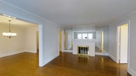 This single-floor space presents a warm and classic interior characterized by rich hardwood flooring that extends throughout the open living area. The focal point of the main room is a white brick fireplace fitted with elegant brass-framed doors, positioned beneath a window that draws in natural light and brightens the surrounding gray-toned walls. Crown molding runs along the ceiling perimeter, adding a refined architectural detail that enhances the traditional character of the space. The layout is notably open, with wide doorways connecting the main living area to adjacent rooms on either side, suggesting a functional and flowing floor plan. The neutral gray and white color palette on the walls provides a clean, versatile backdrop. Doorways visible on both the left and right sides of the fireplace lead to additional rooms, hinting at a well-connected interior arrangement. The space balances classic design elements — such as the brick fireplace and molding details — with a simple, understated aesthetic that lends itself to a variety of interior styles.