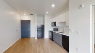 Located at 195 E Main St in the heart of Columbus, Ohio, this single-floor unit offers a clean, modern design with consistent light wood-tone luxury vinyl plank flooring throughout. The open-concept kitchen is outfitted with two-tone cabinetry — white upper cabinets and gray lower cabinets — paired with black appliances including a refrigerator, electric range, over-the-range microwave, and dishwasher. A stainless steel sink and tile backsplash complete the functional and stylish cooking space. The living area is generously proportioned, bathed in natural light from large floor-to-ceiling sliding glass doors that open to an outdoor balcony, offering views of the surrounding Columbus streetscape. Recessed lighting throughout provides consistent, modern illumination. The bedroom is a well-sized, open room with clean white walls and ample space, accessing a walk-in closet visible from the kitchen area. The bathroom features a walk-in shower with a rainfall showerhead, a white single-sink vanity with storage cabinetry, and a full mirror — all finished in a bright, neutral palette. The unit's overall aesthetic is minimalist and contemporary, with a crisp white color scheme and streamlined finishes throughout.