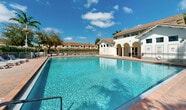 Located at 1140 SE 24th Rd in Homestead, Florida, this outdoor community amenity area centers around a generously sized swimming pool with clear, blue-green water that reflects the South Florida sunshine. The pool is surrounded by a broad brick-paved deck offering ample space for relaxation, lined with multiple lounge chairs and seating arrangements ideal for sunbathing or socializing. A dedicated shallow wading area with convenient step-in access provides a comfortable entry point to the pool. Adjacent to the pool, a charming Mediterranean-style structure features white stucco walls, a terracotta tile roof, and a covered arched loggia, lending a classic Florida aesthetic to the setting. The building appears to serve as a clubhouse or facilities hub for the community. Tall, mature royal palm trees frame the space, enhancing the tropical ambiance and providing natural visual interest against the open sky. The entire pool area is enclosed by a secure metal fence, ensuring safety while maintaining an open, airy feel. Lush green hedges line the perimeter, offering privacy and a verdant backdrop. The surrounding neighborhood is visible in the background, consistent with a well-maintained residential community in Homestead, FL.