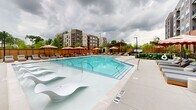Located at 3558 Elm St in Atlanta, GA, this outdoor amenity space is designed to deliver a resort-like experience within a residential community. At its centerpiece is a generously sized swimming pool with crystal-clear turquoise water, featuring a shallow sun shelf with sculptural lounging chairs — ideal for relaxing at the water's edge. The pool deck is finished in light-toned concrete and is lined with rows of cushioned chaise lounges, offering ample space for sunbathing and leisure. Stylish oversized umbrellas in a bold red-and-white stripe pattern provide shaded seating areas outfitted with teak wood furniture and cushioned chairs, perfect for outdoor dining or casual gatherings. Lush landscaping with trees and shrubs is thoughtfully integrated throughout the perimeter, adding privacy and a natural ambiance. A dedicated pet area with black iron fencing, manicured plantings, and pet amenities provides a welcoming space for four-legged residents. The surrounding multi-story building with its mix of brick and modern cladding frames the courtyard, creating an enclosed, community-centered atmosphere. The space is well-appointed, balancing active recreation with relaxed outdoor living.