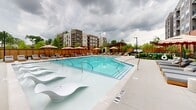 Located at 3558 Elm St in Atlanta, GA, this outdoor amenity space is designed to deliver a resort-like experience within a residential community. At its centerpiece is a generously sized swimming pool with crystal-clear turquoise water, featuring a shallow sun shelf with sculptural lounging chairs — ideal for relaxing at the water's edge. The pool deck is finished in light-toned concrete and is lined with rows of cushioned chaise lounges, offering ample space for sunbathing and leisure. Stylish oversized umbrellas in a bold red-and-white stripe pattern provide shaded seating areas outfitted with teak wood furniture and cushioned chairs, perfect for outdoor dining or casual gatherings. Lush landscaping with trees and shrubs is thoughtfully integrated throughout the perimeter, adding privacy and a natural ambiance. A dedicated pet area with black iron fencing, manicured plantings, and pet amenities provides a welcoming space for four-legged residents. The surrounding multi-story building with its mix of brick and modern cladding frames the courtyard, creating an enclosed, community-centered atmosphere. The space is well-appointed, balancing active recreation with relaxed outdoor living.