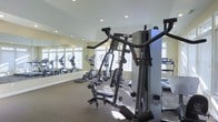 Located at 215 Prairie Creek Way in Columbus, Ohio, this single-floor fitness center is designed to support a well-rounded workout routine. The space is outfitted with a selection of cardio equipment, including multiple elliptical machines positioned along a mirrored wall that visually expands the room and enhances the sense of openness. A prominent multi-function strength training unit anchors the space, offering a variety of resistance-based exercises for users of all fitness levels. The flooring features durable, exercise-appropriate carpeting that provides stability and comfort during workouts. Recessed lighting in the ceiling ensures consistent, even illumination throughout the room, while large windows fitted with blinds allow for adjustable natural light, creating a bright and energizing atmosphere. The neutral color palette of soft greens and whites keeps the environment clean and focused. The layout is thoughtfully organized to allow for safe movement between equipment, making the space functional and accessible. Overall, this fitness center provides a comprehensive and comfortable environment for residents looking to maintain an active lifestyle.