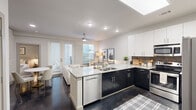 Located in the heart of Dallas, Texas, this single-floor residence at 3015 Routh St offers a thoughtfully designed open-concept layout with contemporary finishes throughout. The kitchen serves as a central focal point, featuring a large granite-topped island with seating, two-tone cabinetry with dark lower cabinets and crisp white upper cabinets, a dark tile backsplash, and a full suite of stainless steel appliances. A skylight above the kitchen floods the space with natural light, complementing the recessed lighting overhead. Dark hardwood-style flooring runs consistently throughout, adding warmth and cohesion to the design. The living and dining area is open and airy, anchored by French doors that lead to an outdoor space, allowing ample natural light to pour in. A ceiling fan overhead and a marble-topped dining table complete the inviting atmosphere. The bedroom is a calm retreat, offering a ceiling fan, natural light from a dedicated window, and direct access to the en-suite bathroom. The bathroom features a long granite vanity with dark cabinetry, a wide mirror, and stylish lighting fixtures. Adjacent to the bedroom is a generous walk-in closet with built-in shelving and hanging rods, providing excellent storage. The overall ambiance is modern, clean, and bright.