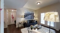 This single-floor residence is situated within a multi-story building that features a stucco exterior, arched entryways, and lush landscaping with palm trees and manicured shrubs. A private balcony with black metal railings offers views of the serene, well-kept courtyard below, adding an outdoor retreat to the living space.

Inside, the living room serves as the central gathering area, showcasing rich hardwood flooring that contrasts elegantly with the neutral, warm-toned walls. A tray ceiling adds architectural depth to the space, while a flush-mount ceiling light provides ambient illumination. The room is appointed with a dark upholstered sofa accented by bold black-and-white patterned throw pillows, flanked by sleek floor lamps that contribute to a contemporary aesthetic. A large black-and-white photographic canvas of a bridge anchors the main wall, lending an urban, artistic character to the room. A glass-top coffee table, a plush white area rug, and thoughtfully placed side tables complete the stylish, well-curated interior. Natural light filters in through a sizable window, brightening the overall ambiance.