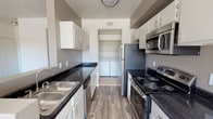 This single-floor residence offers a thoughtfully designed layout with practical features and comfortable living spaces. The galley-style kitchen is a standout, showcasing dark granite countertops, white cabinetry with modern hardware, and stainless steel appliances including a refrigerator, microwave, and electric range. A stainless steel sink with a view down the kitchen corridor adds to the functional design. Wood-look flooring runs throughout, lending a warm and cohesive aesthetic. A dedicated in-unit laundry area is conveniently located just off the kitchen, housing a full-size washer and dryer — a practical amenity that adds everyday convenience. The neutral wall tones and clean finishes create a bright, move-in-ready atmosphere throughout. The private balcony extends the living space outdoors, offering a welcoming retreat with black metal railings and views of a sparkling community pool, well-maintained green lawn, and swaying palm trees. The surrounding Mediterranean-inspired stucco architecture with red tile roofing enhances the resort-like ambiance. The combination of interior functionality and outdoor appeal makes this a well-rounded and inviting space.