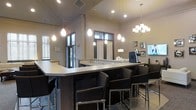 This single-floor communal space is thoughtfully designed to foster a social and inviting atmosphere. At its centerpiece is a large wraparound bar-style counter finished in a sleek, light surface with a dark base, complemented by a row of modern black bar stools that invite guests to gather and relax. The space is illuminated by a combination of elegant teardrop pendant lights and a classic multi-arm chandelier, casting a warm, ambient glow throughout the room. Recessed lighting in the ceiling adds an additional layer of brightness, enhancing the overall sense of openness. The elevated ceilings, accented with architectural molding and detailed trim, lend a refined, upscale character to the interior. Natural light filters in through dark-framed windows and a glass-paneled door, adding depth and connectivity to the outdoors. A coffee or beverage station is visible behind the counter, suggesting the space is well-equipped for hosting and entertaining. Framed artwork adorns the neutral-toned walls, adding personality and warmth. The overall design balances sophistication with comfort, making it an ideal setting for community gatherings, casual meetings, or social events.