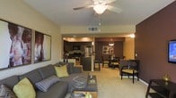 This single-floor residence showcases a thoughtfully designed open-concept floor plan that seamlessly connects the main living areas. The living room serves as the heart of the space, featuring plush gray sectional seating, a sleek glass coffee table, and a ceiling fan with light fixture overhead. Warm beige carpeting flows throughout, complementing the neutral wall tones accented by a rich chocolate-brown feature wall that visually anchors the space. A piece of large-scale artwork adds a bold decorative touch to the living area.

The kitchen is equipped with stainless steel appliances, including a refrigerator and range, and features dark cabinetry paired with a center island or peninsula that provides additional prep and seating space. Pendant lighting adds a refined touch above the kitchen area.

Adjacent to the kitchen, the dining area accommodates a full dining set with upholstered chairs, lit by a stylish pendant chandelier. The cohesive color palette of warm taupes, deep browns, and green accent tones ties all areas together, creating a welcoming and sophisticated ambiance throughout. The open layout maximizes the sense of space and allows for easy flow between entertaining and everyday living areas.