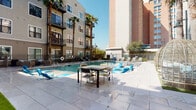 This outdoor amenity space is designed to deliver a resort-style experience within a modern multi-story residential community. At its centerpiece is a generously sized swimming pool with clear blue water, flanked by a broad, light-toned stone tile sun deck that provides ample room for lounging and socializing. Outdoor dining tables and chairs are arranged poolside, offering a comfortable setting for gatherings under open skies.

Surrounding the pool area, vibrant turquoise chaise lounges and accent furniture add pops of color against the neutral hardscape. A dedicated turf lawn area with additional lounge chairs creates a softer, relaxed retreat, ideal for sunbathing away from the pool's edge. Tall palm trees punctuate the space, lending a warm, resort-inspired aesthetic and providing natural shade.

A fully equipped outdoor kitchen station features a large stainless steel built-in grill and a tiled counter surface, making it an ideal spot for al fresco cooking and entertaining. A sculptural woven pod chair adds a distinctive design touch to one corner of the space.

The entire area is enclosed by sleek black metal fencing with gated access points, ensuring privacy and security. The multi-story building façade, complete with balconies overlooking the courtyard, frames the space and contributes to its intimate, community-centered atmosphere.