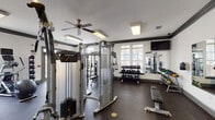 Located at 26 Anvil Peak Dr in Garner, NC, this dedicated fitness center offers a comprehensive workout environment designed to accommodate a wide range of exercise routines. The space is finished with durable rubber flooring throughout and features crisp white walls accented by dark crown molding, giving the room a clean, structured aesthetic. Ceiling fans provide consistent air circulation, complemented by recessed and flush-mounted lighting that keeps the facility well-lit at all times. Large windows along the perimeter walls allow natural light to fill the space while offering views of the surrounding community. The cardio section is well-appointed with multiple Precor treadmills, elliptical trainers, and a stationary bike, each equipped with individual monitors. The strength training area includes a Precor cable machine system, a multi-use functional trainer, and additional selectorized equipment for targeted muscle training. A tiered dumbbell rack holds a full range of free weights, while a dedicated medicine ball rack provides options for functional fitness and conditioning work. Wall-mounted mirrors on multiple sides enhance the sense of space and allow users to monitor their form. Wall-mounted televisions are positioned for visibility from the cardio equipment, adding to the overall user experience of this fully outfitted fitness facility.