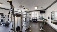 Located at 26 Anvil Peak Dr in Garner, NC, this dedicated fitness center offers a comprehensive workout environment designed to accommodate a wide range of exercise routines. The space is finished with durable rubber flooring throughout and features crisp white walls accented by dark crown molding, giving the room a clean, structured aesthetic. Ceiling fans provide consistent air circulation, complemented by recessed and flush-mounted lighting that keeps the facility well-lit at all times. Large windows along the perimeter walls allow natural light to fill the space while offering views of the surrounding community. The cardio section is well-appointed with multiple Precor treadmills, elliptical trainers, and a stationary bike, each equipped with individual monitors. The strength training area includes a Precor cable machine system, a multi-use functional trainer, and additional selectorized equipment for targeted muscle training. A tiered dumbbell rack holds a full range of free weights, while a dedicated medicine ball rack provides options for functional fitness and conditioning work. Wall-mounted mirrors on multiple sides enhance the sense of space and allow users to monitor their form. Wall-mounted televisions are positioned for visibility from the cardio equipment, adding to the overall user experience of this fully outfitted fitness facility.
