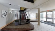 This interior common area presents a well-appointed lobby space defined by a central open-riser staircase constructed with dark metal railings and wood-toned treads. The staircase sits atop a raised, brick-bordered platform finished with exposed aggregate, lending the feature a sculptural, focal quality. Flanking the staircase are tall, slender decorative branches and ornamental grasses arranged in square planters, adding an organic contrast to the structured architectural elements. The flooring transitions between light-toned ceramic tile and the textured aggregate platform, creating clear zones within the space. A substantial cylindrical column anchors one side of the room, reinforcing the building's structural character. Along the far wall, a row of what appear to be individual storage lockers or mailboxes lines the corridor, suggesting a functional residential or mixed-use facility. Wall-mounted sconce lighting supplements the ambient illumination, while large glazed panels along the perimeter allow natural light to fill the space. The textured ceiling and neutral wall tones contribute to a clean, utilitarian aesthetic throughout.