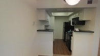This single-floor space presents a practical and well-organized layout centered around a compact yet functional kitchen. The kitchen is equipped with black appliances, including a refrigerator, gas range, and over-the-range microwave, all complemented by crisp white cabinetry that provides ample storage. Dark countertops offer a clean contrast against the white cabinets, lending a classic, understated aesthetic to the space. A peninsula with a dark countertop surface extends from the kitchen, creating a natural division between the cooking area and the adjacent space, while also offering additional workspace or casual seating potential. The flooring transitions from wood-look planks in the kitchen to a lighter surface in the connecting area, adding visual interest throughout. A recessed ceiling detail above the kitchen adds architectural dimension, while the light-toned walls keep the overall atmosphere bright and open. The layout is designed to maximize efficiency, making thoughtful use of the available space. Overall, the space offers a functional and neatly appointed interior with cohesive design elements throughout.