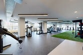 Located at 325 15th St in Jersey City, NJ, this state-of-the-art fitness center offers a generously proportioned, single-floor layout designed to accommodate a full range of athletic and training activities. The space is thoughtfully organized into distinct workout zones, each catering to different fitness disciplines.

The cardio area features a row of modern treadmills and stair climbers, all equipped with individual screens, positioned along large windows that flood the space with natural light and provide views of the surrounding neighborhood. A dedicated strength training section hosts a comprehensive selection of cable machines, weight benches, plate-loaded equipment, and a dumbbell rack, offering everything needed for a complete resistance workout.

A standout feature is the artificial turf training strip, ideal for functional fitness, sled drags, agility drills, and dynamic movement exercises. The flooring throughout the remainder of the space consists of durable rubber-speckled material, providing both safety and comfort underfoot.

The interior design is clean and modern, with white walls, recessed lighting, and sleek linear pendant fixtures running across the ceiling. Exposed industrial ductwork adds a contemporary edge to the aesthetic. Full-length mirrors along one wall enhance the sense of openness. The overall ambiance is bright, professional, and well-suited for serious training in an accessible urban setting.