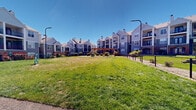 Located at 9770 Rosemont Ave in Lone Tree, Colorado, this outdoor community amenity space serves as a dedicated dog park and recreational green area set against the backdrop of a well-maintained multi-story residential complex. The grounds feature a generous expanse of open, manicured lawn that provides ample room for pets and residents to enjoy the Colorado outdoors under clear blue skies.

A central wooden pergola serves as a focal point of the space, offering a shaded seating area equipped with a sturdy metal picnic table and benches — ideal for relaxing while supervising pets. The dog park area is fully enclosed with chain-link fencing, ensuring a safe and secure environment, and includes a designated entry gate with clear access pathways of compacted gravel.

Pet agility equipment, including tire jump obstacles, is positioned throughout the lawn area to provide enrichment and activity for dogs of all sizes. Mature trees and neatly trimmed shrubs are landscaped along the perimeter, adding natural beauty and privacy to the space. Tall street lamps are placed throughout, supporting usability during early morning and evening hours.

The surrounding residential buildings feature multi-level balconies, arched windows, brick accents, and pitched rooflines, giving the community a cohesive and attractive architectural character. The entire space is thoughtfully designed to foster outdoor activity and community engagement in the heart of Lone Tree.