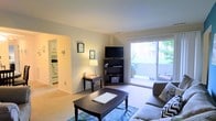 This single-floor residence features a welcoming living room as its central gathering space, finished with wall-to-wall carpeting and neutral-toned walls that create a warm, understated ambiance. The room is well-lit, benefiting from abundant natural light streaming through a large sliding glass door that opens directly onto an outdoor patio, offering pleasant views of surrounding greenery. The patio extends the living space outdoors, adding a functional area for relaxation.

The living room is thoughtfully arranged with room for a sofa, coffee table, and entertainment setup, including a corner bookshelf and wall-mounted media unit. Framed wall art and a table lamp add decorative character to the space. A hallway visible from the living area connects to the kitchen, which can be partially seen and appears to include standard appliances and cabinetry, suggesting a functional and efficient layout. The open sightlines between the living room and kitchen contribute to a cohesive, connected feel throughout the home. Overall, the space presents a comfortable and practical single-level layout with a bright, airy atmosphere.