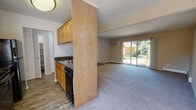 This single-floor, two-bedroom, one-bathroom Alpine unit is thoughtfully laid out to maximize functionality and comfort. The galley-style kitchen features warm honey-toned wood cabinetry lining both walls, dark countertops, and black appliances including a range, refrigerator, and dishwasher, all set atop durable wood-look flooring. A half-wall partition separates the kitchen from the adjacent living area, creating a sense of openness while maintaining definition between spaces. The spacious living room is carpeted and bathed in natural light, anchored by a large sliding glass door that opens to a view of lush greenery and mature trees, giving the space an inviting, airy feel. A wall-unit air conditioner and baseboard heating ensure year-round comfort. The bedrooms are carpeted and well-proportioned, each offering windows that frame peaceful outdoor scenery and baseboard heating. The bathroom is clean and functional, featuring a wood-toned vanity with dark countertop, a large mirror, tile flooring, and a shower-tub combo enclosed by sliding glass doors. A closet-lined hallway connects the main living areas to the bedrooms and bathroom, providing ample storage throughout this well-organized unit.