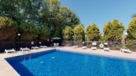 This outdoor pool area offers a refreshing and well-maintained retreat designed for relaxation and recreation. At its center sits a generously sized swimming pool with crystal-clear blue water, accessible via built-in entry steps and equipped with safety features including a life ring station. The surrounding concrete sun deck provides ample space for lounging, lined with white reclining chairs positioned to take full advantage of open sky and natural sunlight.

Shaded gathering areas are thoughtfully distributed around the deck, featuring umbrella-covered tables and chairs — ideal for casual outdoor dining or socializing. The pool area is enclosed by a black wrought iron fence, offering both security and an unobstructed view of the lush, mature trees that line the perimeter, providing a natural and serene backdrop.

Adjacent to the pool, a brick building with large French doors offers convenient access to the pool deck, along with additional outdoor seating and amenities. Pole-mounted lights are positioned throughout the space, extending the usability of the area into the evening hours. The overall ambiance is bright, open, and inviting, combining practical amenities with a pleasant outdoor setting suited for warm-weather enjoyment.
