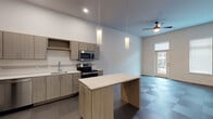 This single-story space in Rogers, Arkansas, showcases a clean, modern design with thoughtful finishes throughout. The kitchen serves as a central highlight, outfitted with light wood-grain cabinetry, white quartz countertops, and a full suite of stainless steel appliances including a refrigerator, electric range, microwave, and dishwasher. A freestanding island with a matching white surface provides additional workspace and casual seating. Elegant cylindrical pendant lights hang above the island, while patterned tile flooring adds visual interest underfoot.

The open living area flows naturally from the kitchen, featuring a ceiling fan, recessed lighting, and access to a windowed exterior door that invites natural light. Bedrooms are carpeted and benefit from notably high ceilings, creating an airy, spacious feel. Tall, narrow windows fitted with white blinds provide soft, diffused light. Each bedroom also connects to adjacent spaces, including a walk-in closet area.

The bathroom continues the modern aesthetic with a wide dual-sink vanity in light wood tones, white quartz countertop, rectangular undermount sinks, chrome hardware, and two large framed mirrors. A separate shower with tiled surround completes the space. Overall, the unit maintains a consistent, contemporary palette throughout.