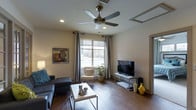 This 1-bedroom, 1-bathroom unit offers a well-designed single-floor layout that balances comfort and functionality. The living area serves as the heart of the space, featuring rich hardwood-style flooring that extends throughout and lends a warm, inviting ambiance. A large window floods the room with natural light while framing an outdoor view, complemented by elegant grey curtains that add a touch of sophistication. A stylish ceiling fan with a brushed metal finish is centrally mounted, providing both practical airflow and a decorative accent. Recessed lighting adds to the well-lit atmosphere of the room. The living space is thoughtfully arranged to accommodate a sofa, accent chair, and a media console, making it ideal for relaxing and entertaining. Neutral wall tones create a versatile backdrop that pairs well with the contemporary furnishings. A framed piece of wall art adds a colorful focal point to the space. The overall design aesthetic is modern and clean, with attention to detail evident in the coordinated decor and finishes. This unit provides a comfortable and stylish living environment suited for everyday living.